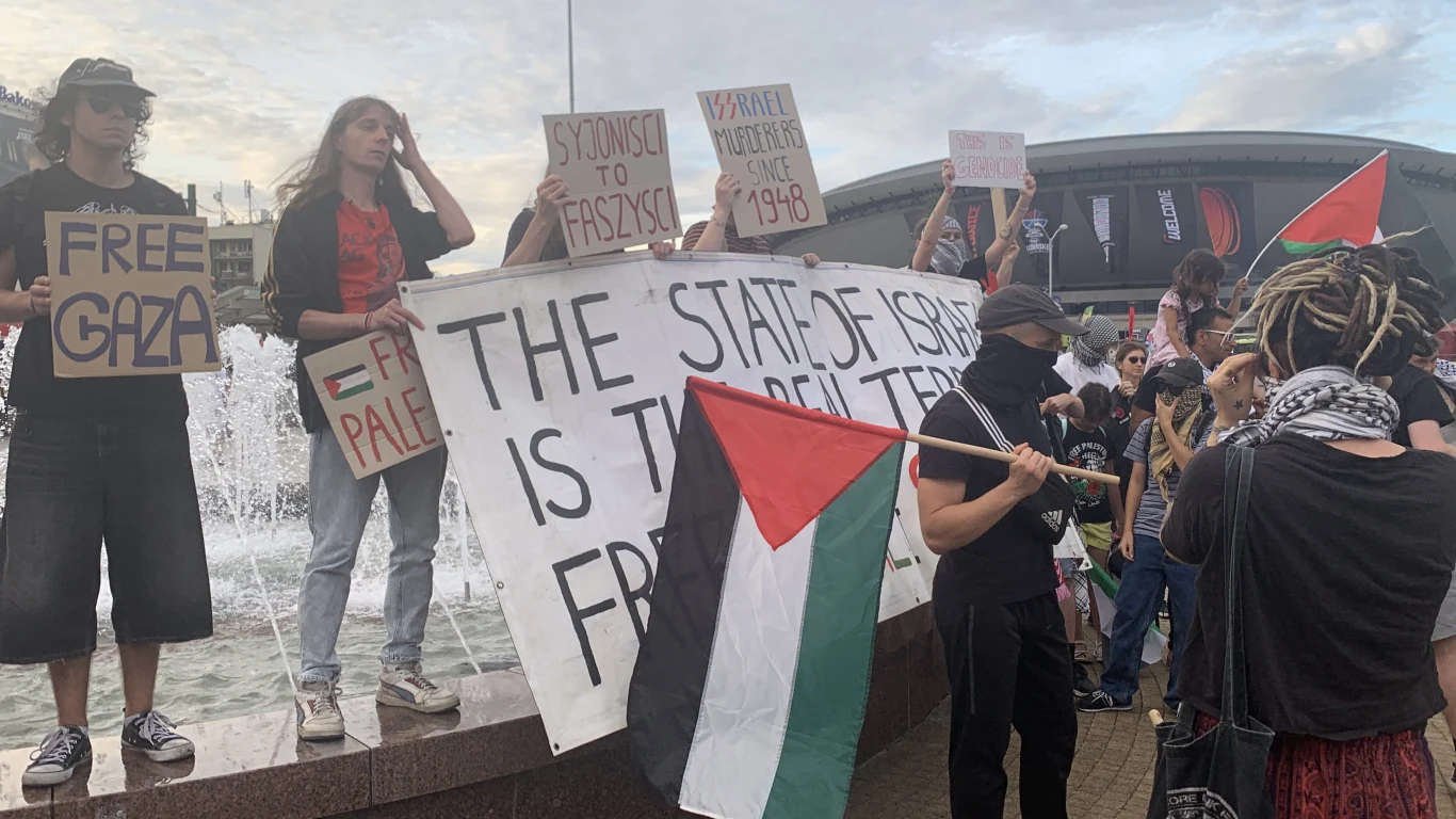 Grupa osób uczestniczących w demonstracji z transparentami oraz flagą Palestyny, trzymających plakaty z hasłami politycznymi dotyczącymi konfliktu izraelsko-palestyńskiego, tłum zgromadzony przy fontannie na tle miejskiej architektury.