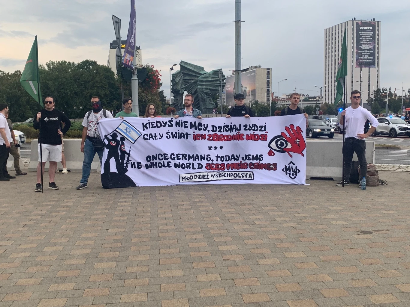 Grupa osób stojących na miejskim placu, trzymających transparent z hasłami antysemickimi napisanymi po polsku i angielsku, w tle widoczne budynki i zielone flagi.