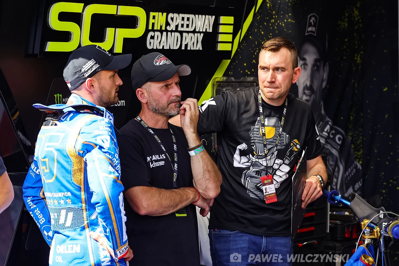 Bartosz Zmarzlik we GP Polski we Wrocławiu korzystał ze wsparcia tunera Daniela Kowalskiego. Trzech mężczyzn rozmawia w boksie podczas zawodów FIM Speedway Grand Prix, jeden ubrany w strój żużlowca, pozostali noszą czarne koszulki i identyfikatory, na tle wyeksponowane są logotypy oraz tło związane ze sportem motorowym.