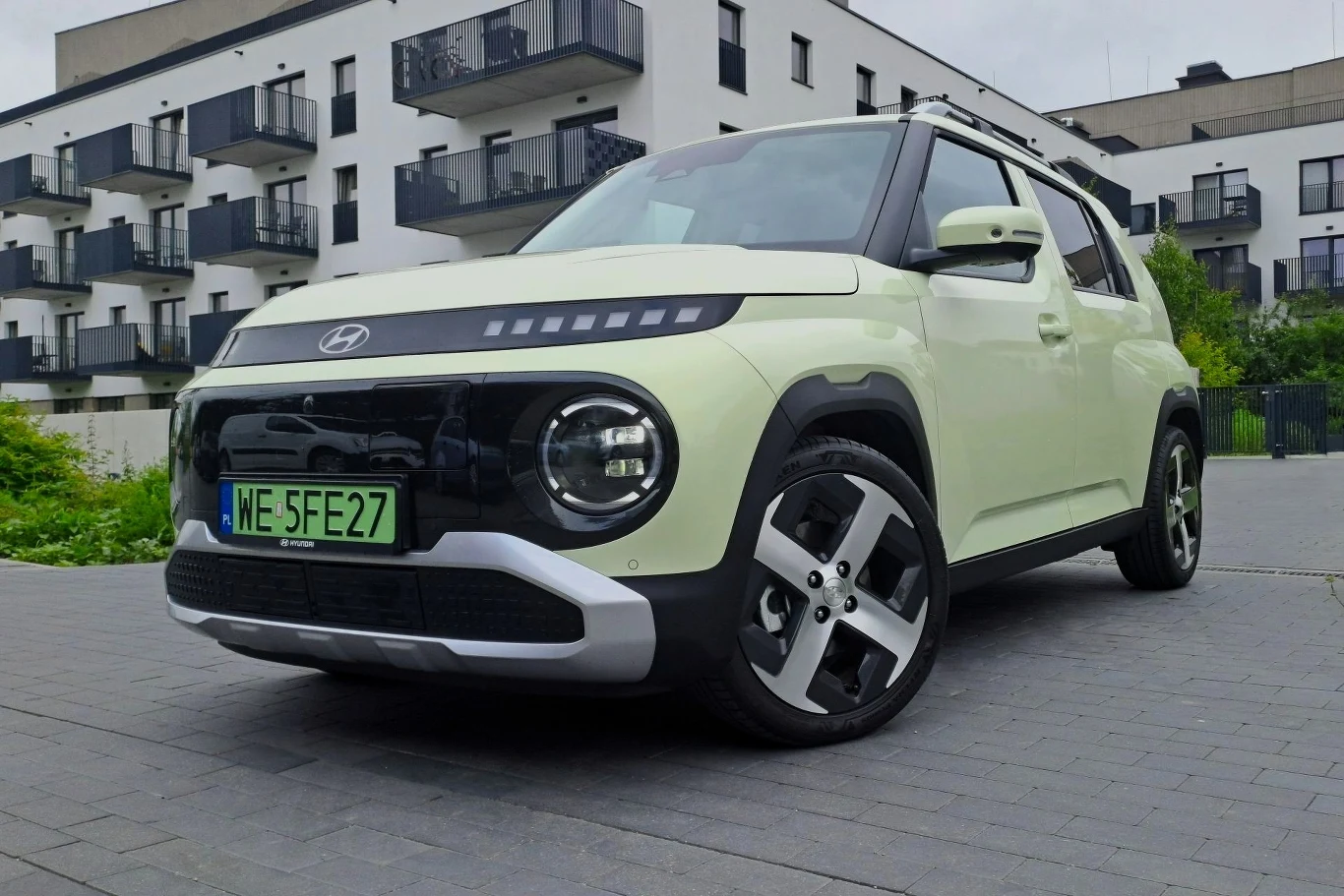 Hyundai Inster to kolejny stosunkowo tani elektryk na rynku. Czy zabierze klientów Dacii Spring? Nowoczesny samochód Hyundai o jasnej karoserii z czarnymi akcentami stoi zaparkowany na tle nowoczesnych budynków mieszkalnych, na przednim planie widoczna stylowa felga i zielona tablica rejestracyjna.