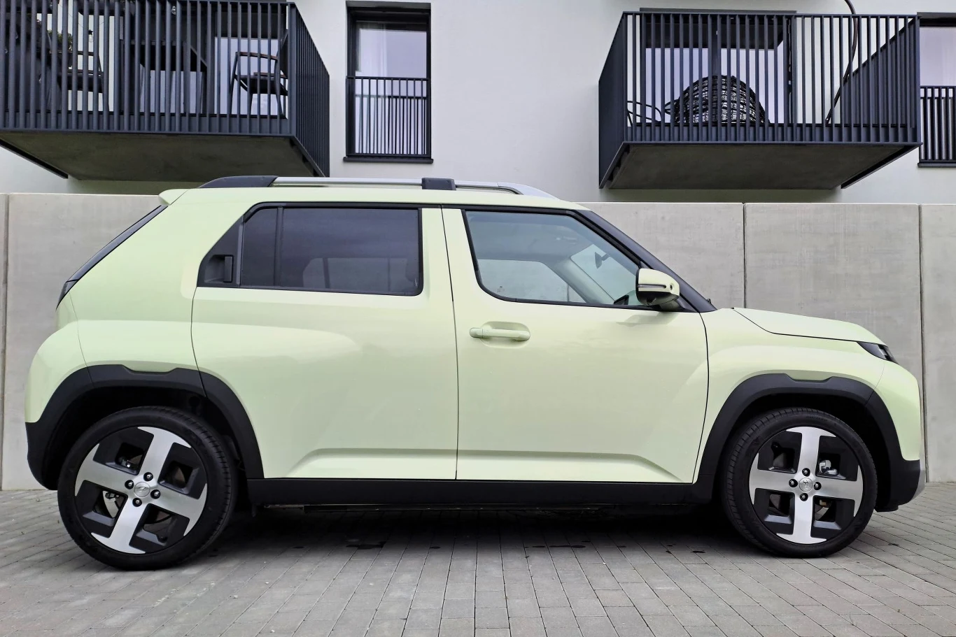 Hyundai Inster ma o 4 mm większą wysokość względem szerokości. Jasnozielony samochód terenowy o nowoczesnej sylwetce zaparkowany na kostce brukowej przed szarym murem, w tle widoczne balkony współczesnego budynku mieszkalnego.