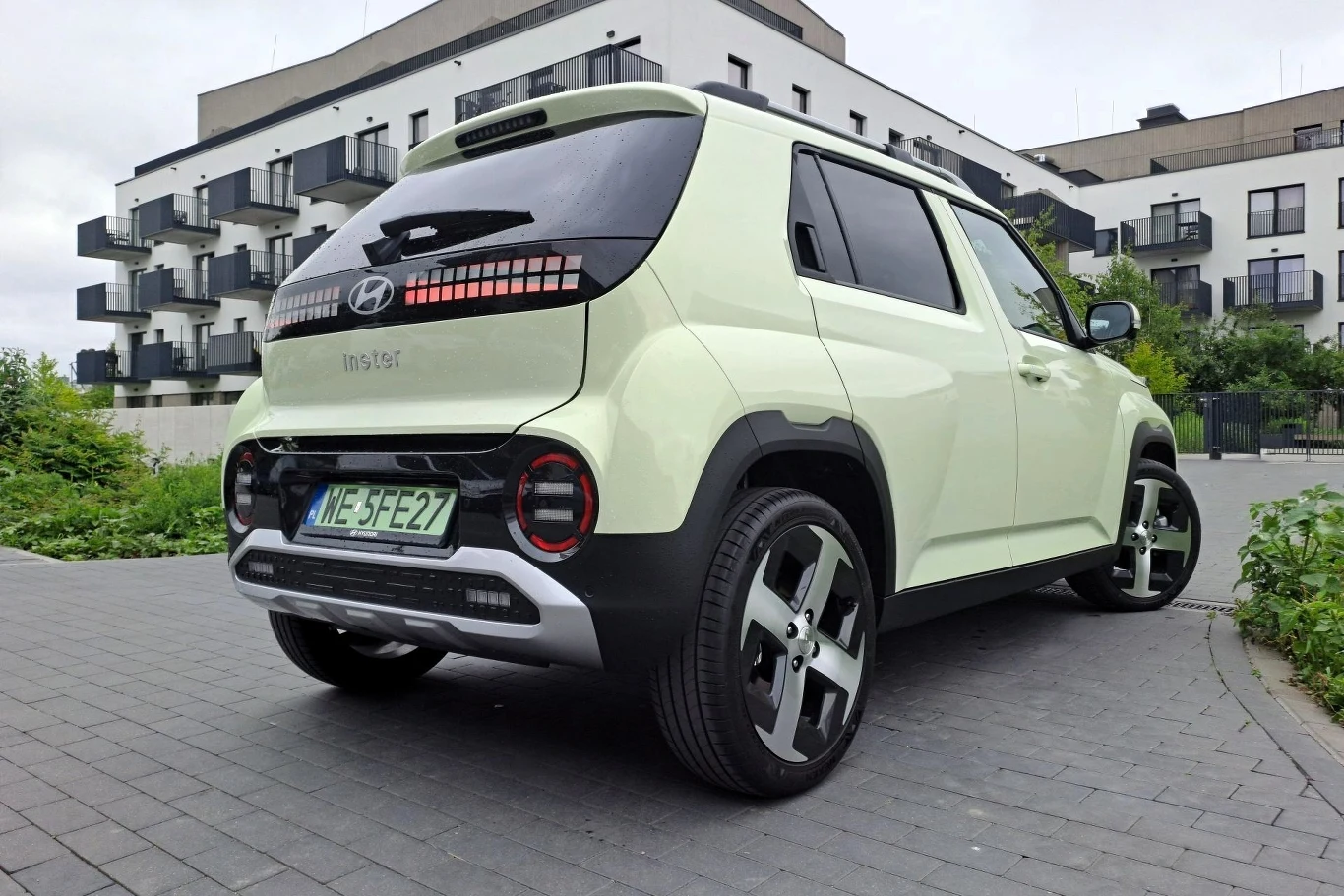 Dzięki systemowi rekuperacji z aktywowaną funkcją i-Pedal praktycznie wcale nie musimy korzystać z hamulca. Mały, miejski samochód Hyundai o białym kolorze zaparkowany na nowoczesnym osiedlu mieszkaniowym, w tle widać białe budynki z balkonami i zielenią.