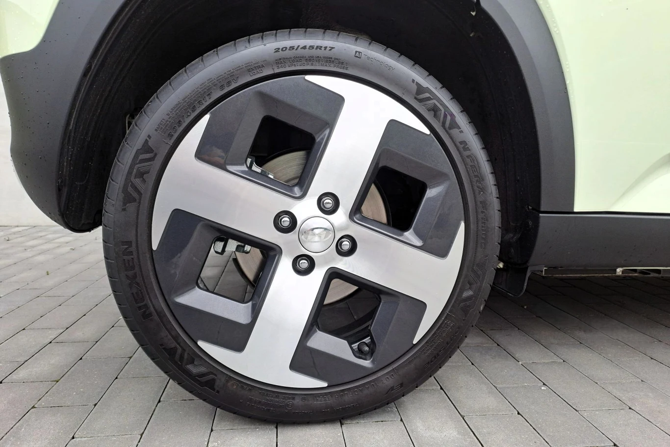 Hyundai Inster dostępny jest z 15-calowymi felgami stalowymi oraz felgami aluminiowymi w rozmiarach 15 lub 17 cali. Koło samochodowe z charakterystyczną felgą aluminiową o matowo-czarno-srebrnym wykończeniu, zamontowane na pojeździe zaparkowanym na kostce brukowej.