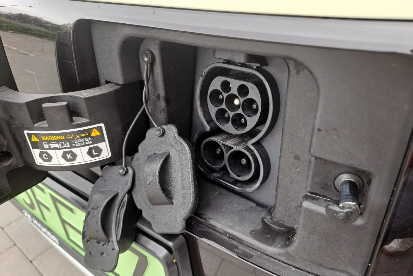 W zależności od specyfikacji Hyundai Inster może przejechać do 370 km na jednym ładowaniu. Gniazdo ładowania do samochodu elektrycznego z otwartymi klapkami zabezpieczającymi, widoczne etykiety ostrzegawcze i złącza do ładowania na zewnętrznej obudowie pojazdu.