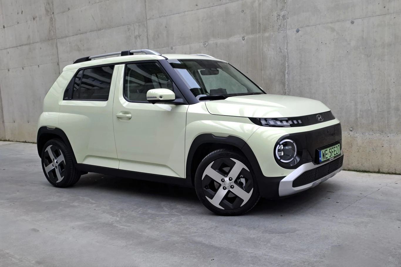 W ramach akcji promocyjnej ceny Hyundaia Instera startują obecnie od 101 900 zł (cena katalogowa: 107 900 zł). Nowoczesny, jasny samochód SUV o kompaktowych rozmiarach, zaparkowany przy betonowej ścianie, wyróżniający się prostym, geometrycznym designem nadwozia, czarnymi elementami wykończenia oraz charakterystycznymi felgami.