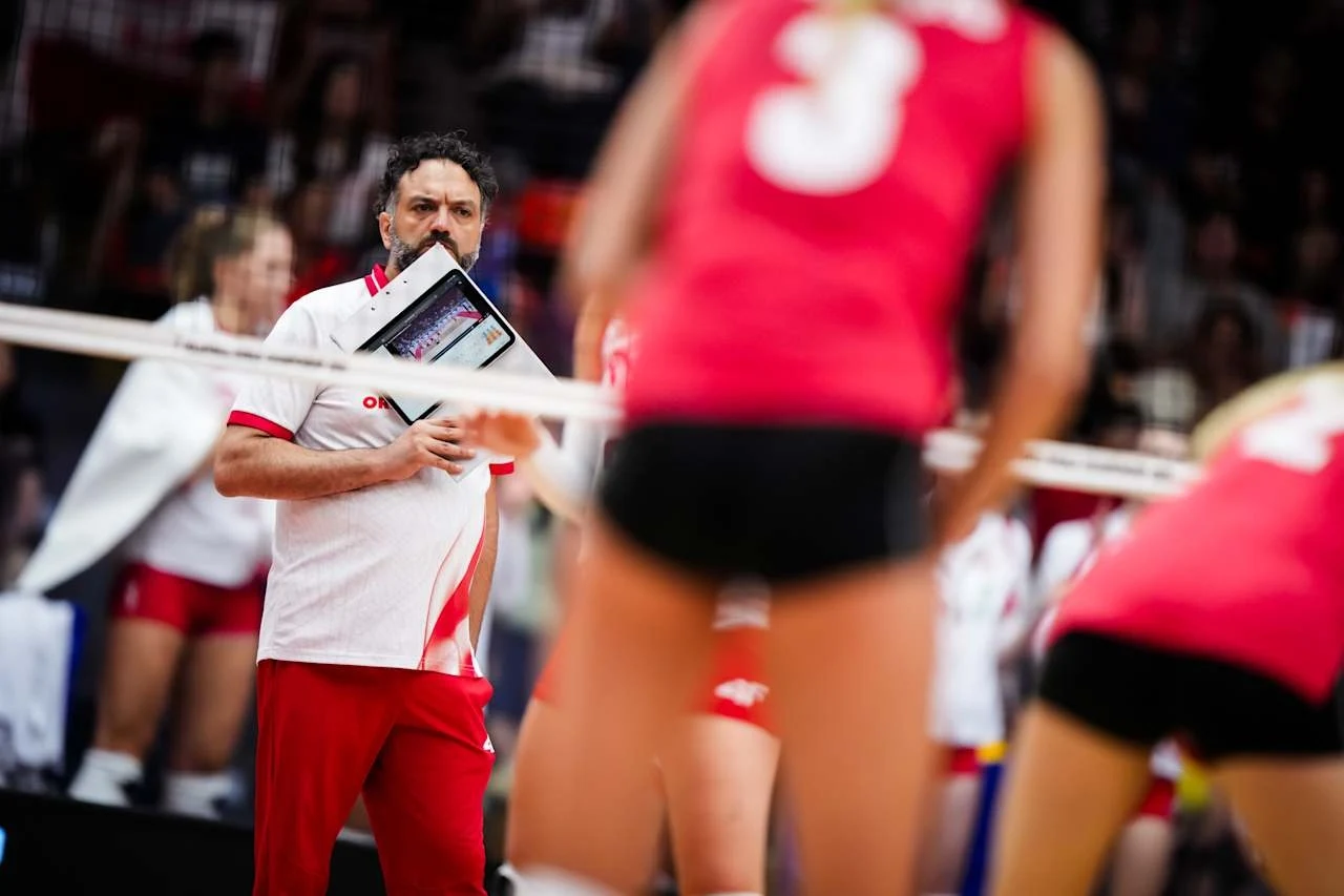 Trener w białej koszulce z czerwoną lamówką i czerwonych spodniach trzyma tablicę taktyczną przy twarzy, patrząc na boisko siatkarskie podczas meczu; na pierwszym planie siatkarki w czerwonych strojach przygotowujące się do zagrania.