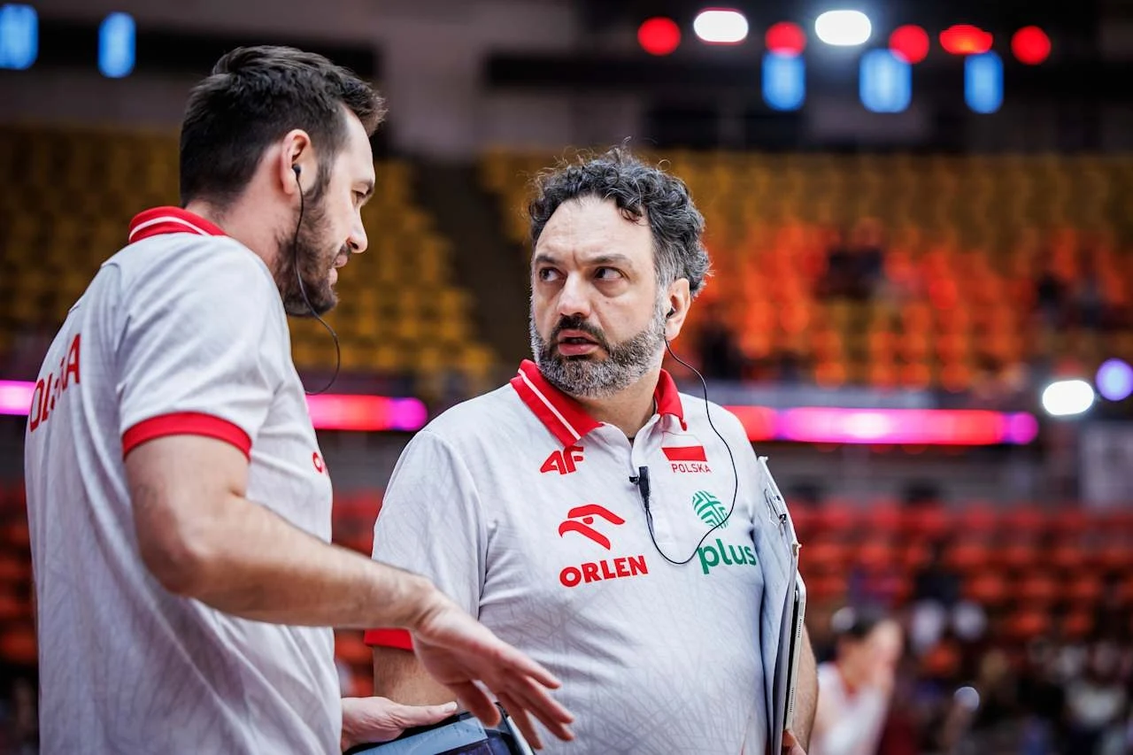 Dwóch mężczyzn w sportowych koszulkach z polskimi oznaczeniami rozmawia ze sobą na tle trybun hali sportowej, jeden trzyma w ręku notatnik, obaj skupieni na rozmowie.