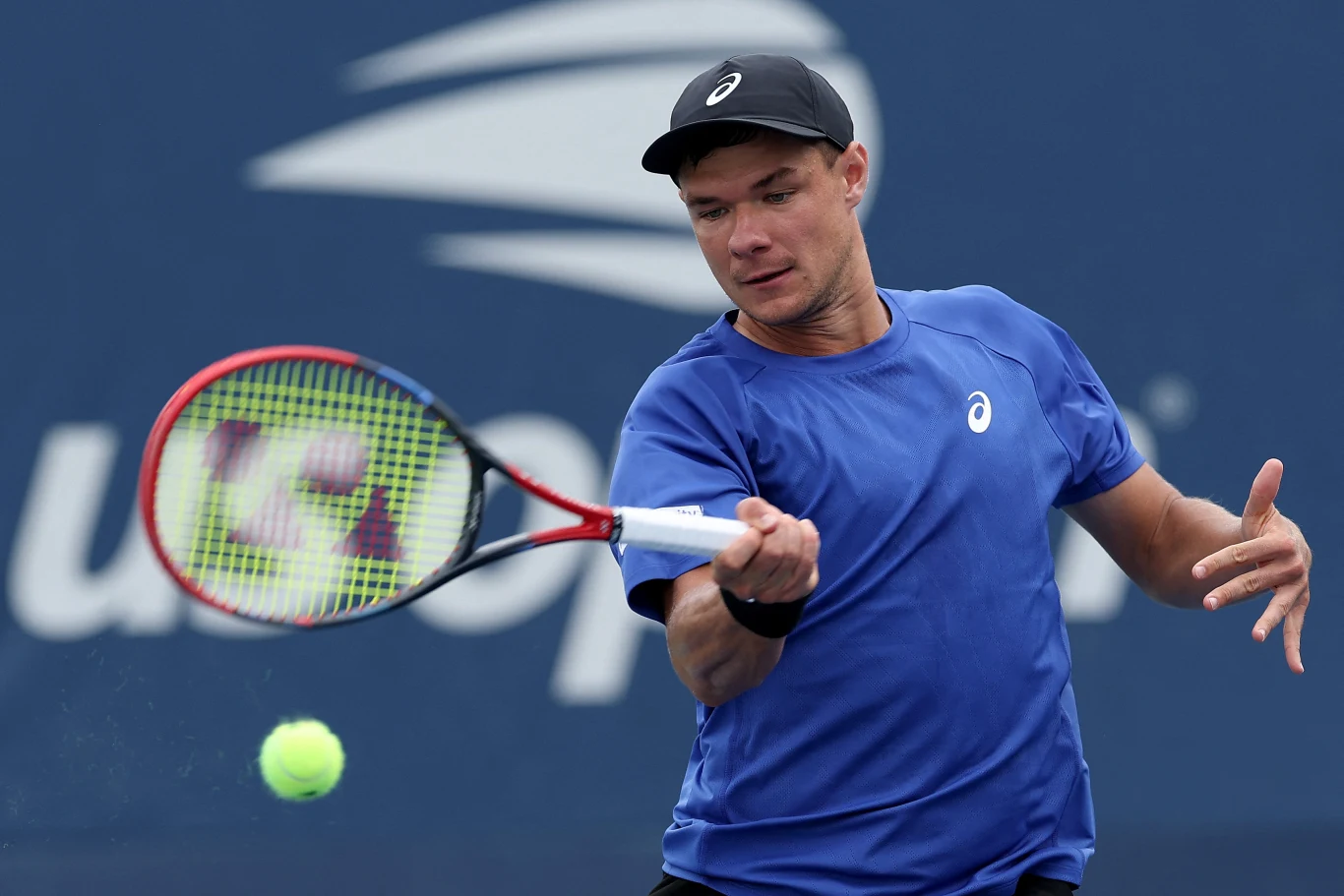 Tenisista w niebieskiej koszulce uderza piłkę rakietą podczas meczu na tle niebieskiego kortu tenisowego.