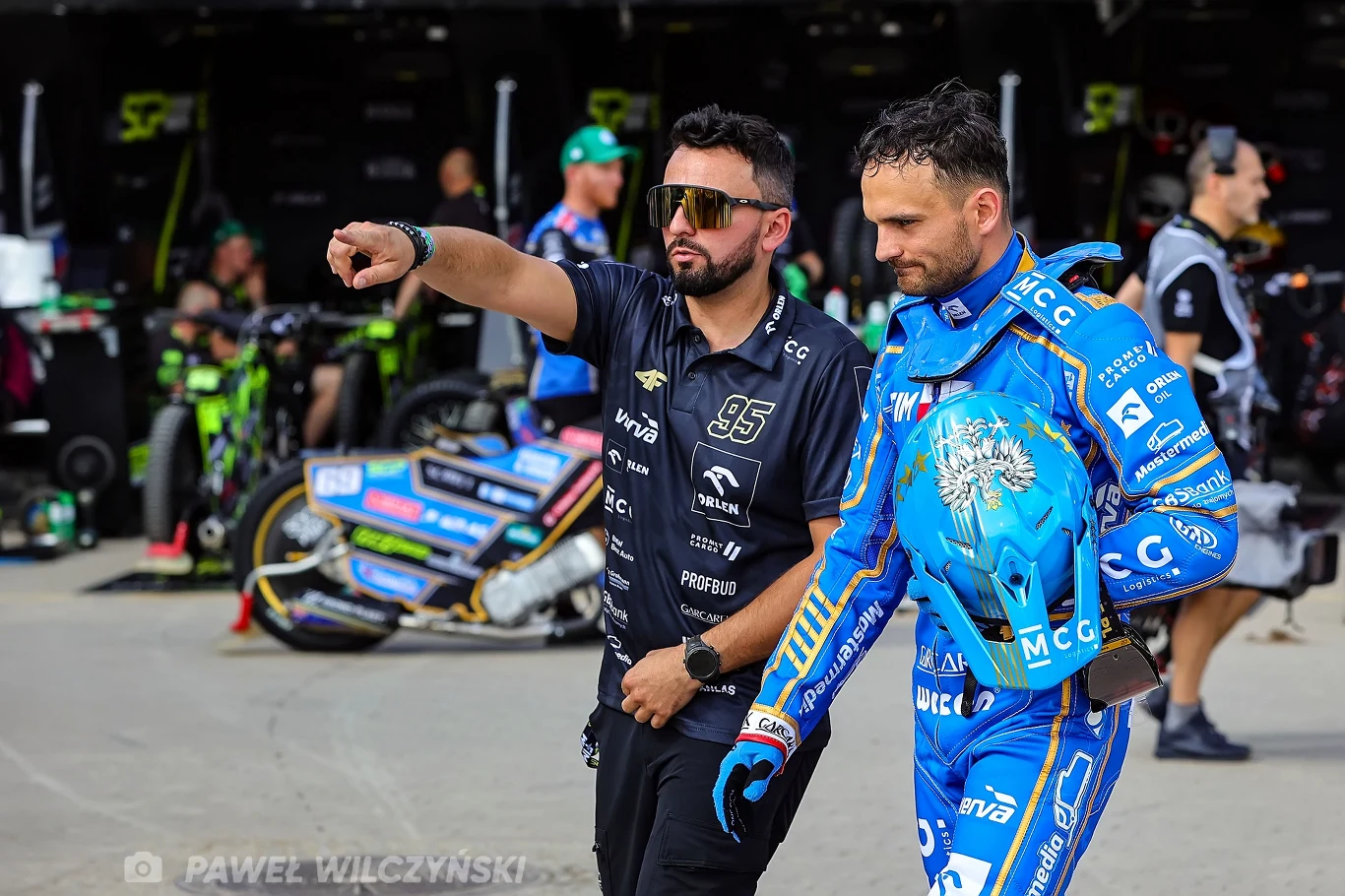 Paweł Zmarzlik w dyskusji z Bartoszem Zmarzlikiem Dwóch mężczyzn znajduje się w strefie serwisowej podczas wydarzenia sportowego, jeden z nich w stroju wyścigowym trzyma kask, drugi wskazuje coś poza kadrem; w tle motocykle i obsługa techniczna.
