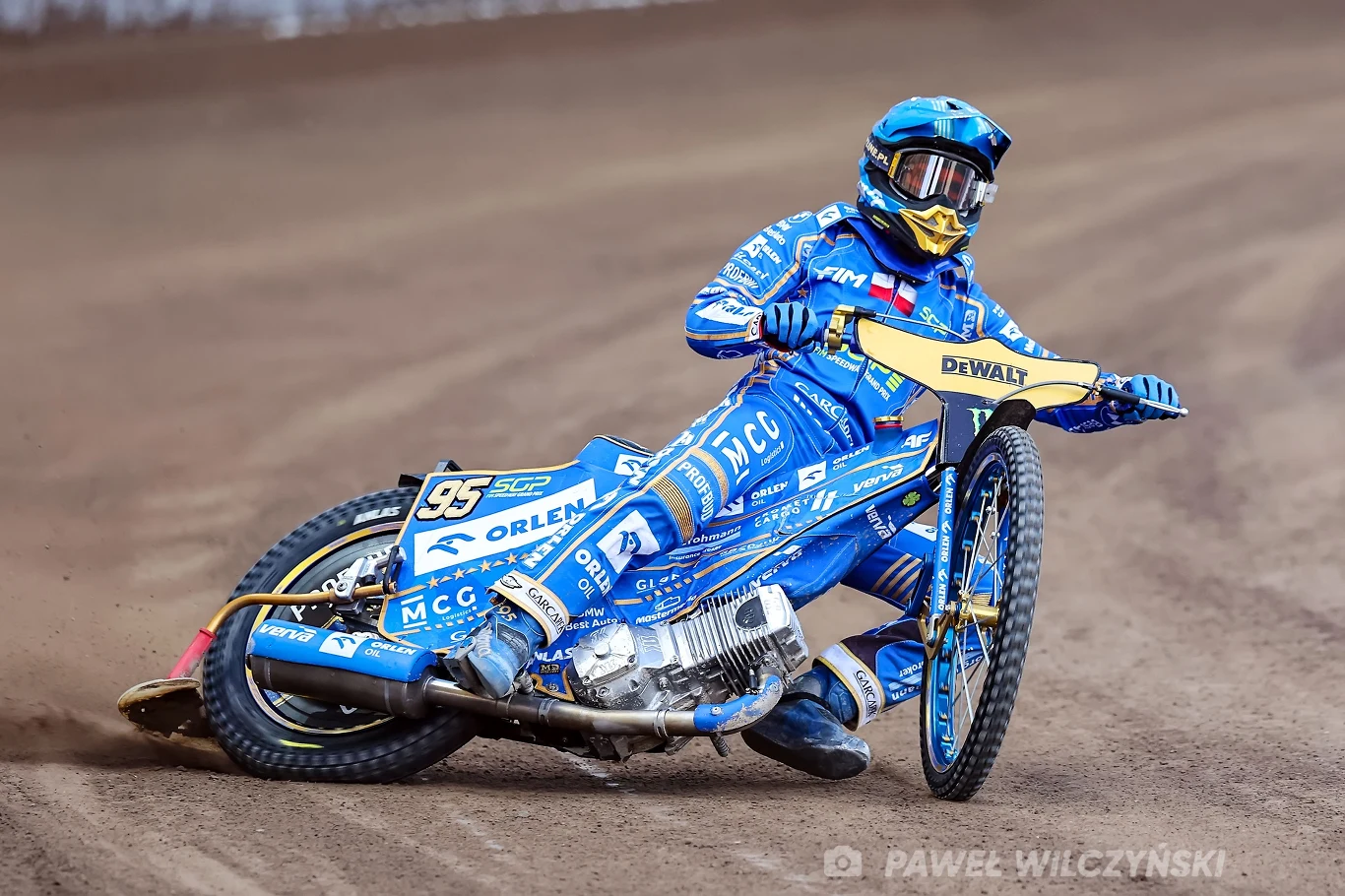 Zawodnik żużlowy w niebieskim kombinezonie i kasku pochylony podczas dynamicznego skrętu na motocyklu na torze żużlowym, widoczna szczegółowa grafika motocykla z logo sponsorów.