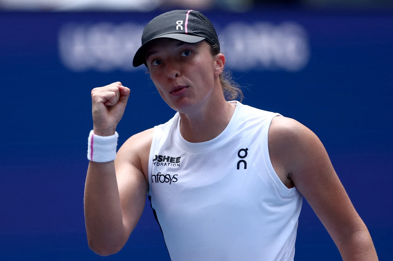 Tennistka w sportowej koszulce i czapce z daszkiem unosi zaciśniętą pięść w geście zwycięstwa lub motywacji, skoncentrowana, na tle zamazanego napisu turniejowego.