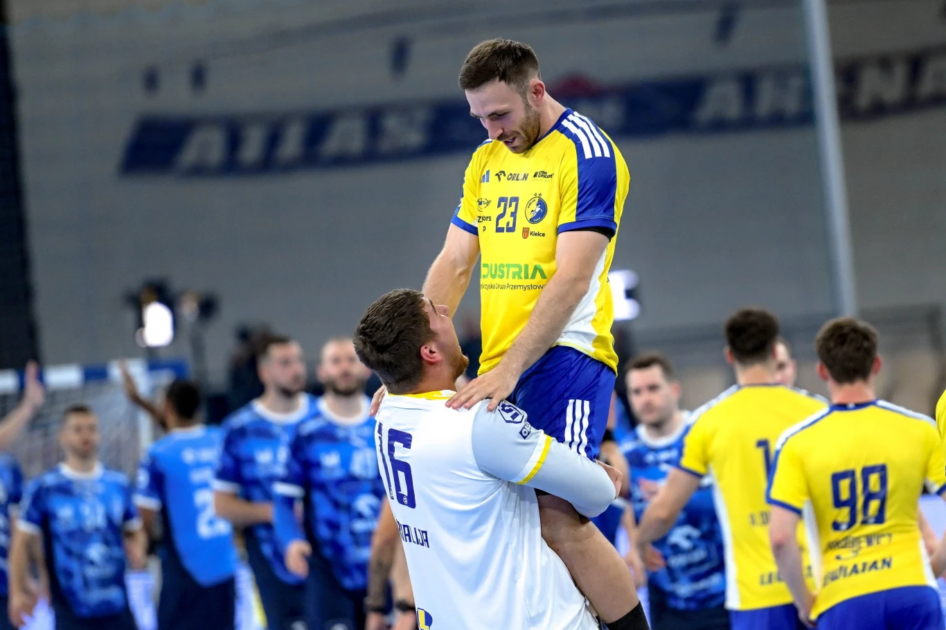 Zawodnik w żółto-niebieskim stroju uniesiony w górę przez współzawodnika w białej koszulce podczas meczu piłki ręcznej, w tle drużyna ubrana w niebieskie stroje oraz hala sportowa.