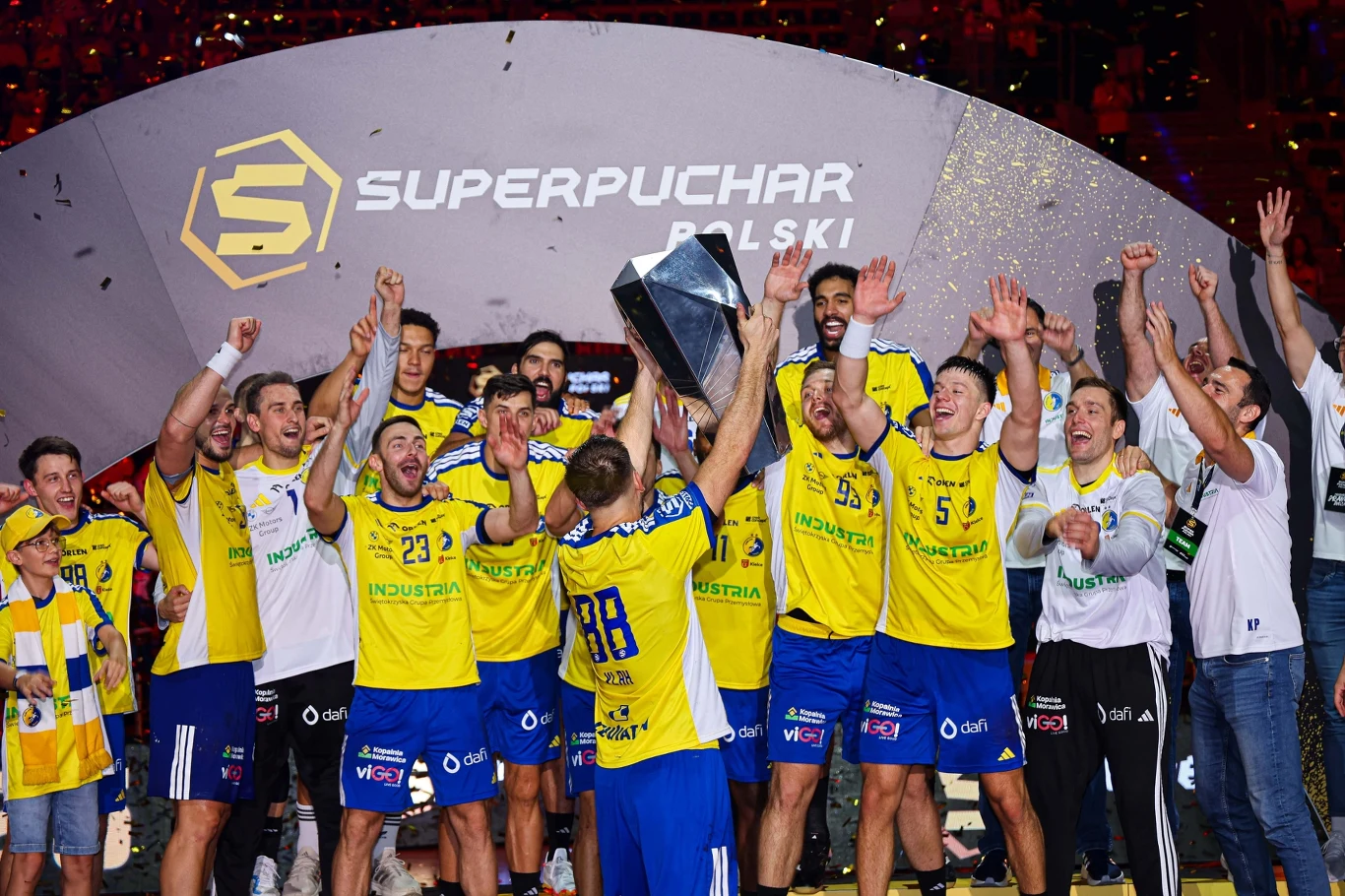 Zawodnicy drużyny piłki ręcznej ubrani w żółto-niebieskie stroje świętują zwycięstwo, trzymając trofeum Superpucharu Polski, nad nimi konfetti, w tle logo wydarzenia.