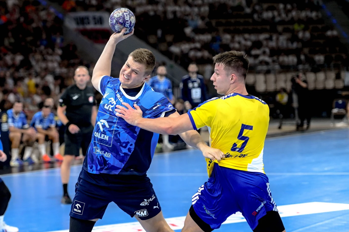 Dwóch zawodników walczy o piłkę podczas meczu piłki ręcznej, jeden w niebieskim stroju z numerem 22 unosi piłkę nad głową, drugi w żółto-niebieskim stroju z numerem 5 blokuje przeciwnika ręką, w tle widoczna publiczność oraz sędzia.