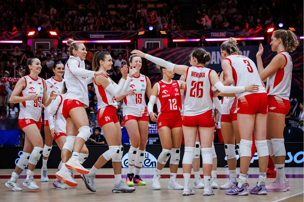 Reprezentacja Polski w siatkówce kobiet celebrująca punkt podczas meczu, zawodniczki ubrane w stroje sportowe radośnie przybijają sobie piątki, w tle widoczna publiczność.