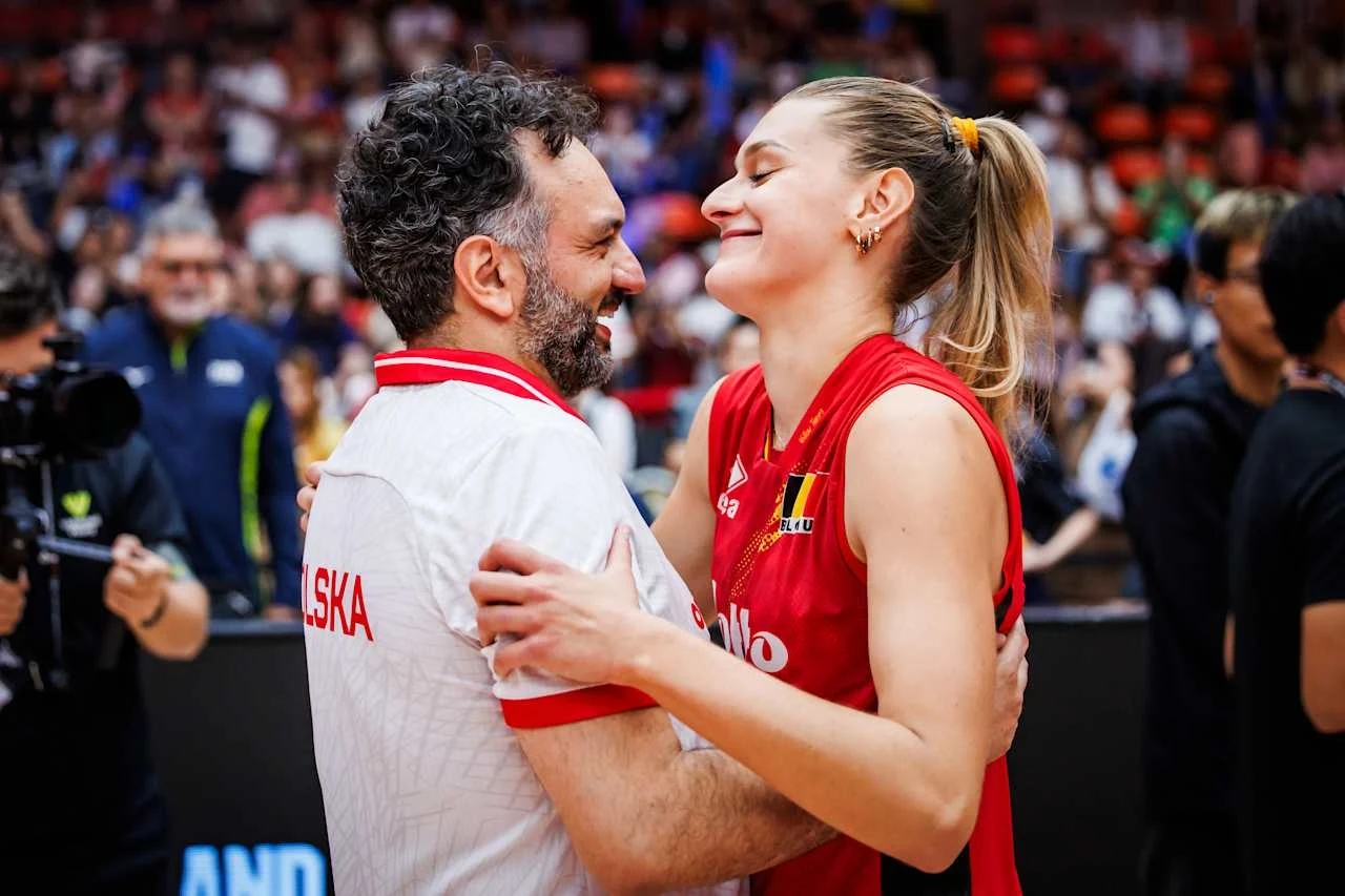 Dwójka uśmiechniętych sportowców obejmuje się na tle widowni, mężczyzna w białej koszulce z napisem 'Polska', kobieta w czerwonym stroju sportowym z emblematem, atmosfera radości i gratulacji.