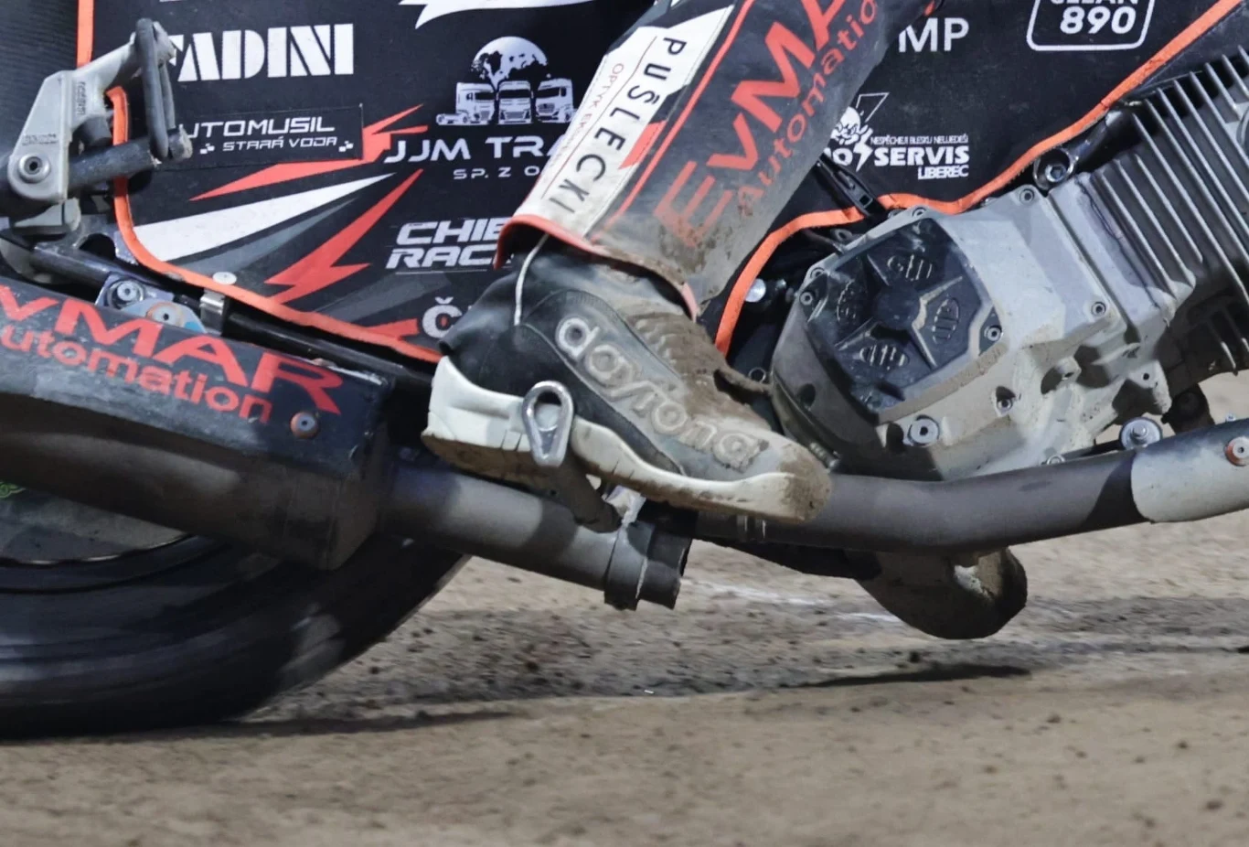 Tłumik Jana Kvecha Motocykl wyścigowy podczas dynamicznej jazdy po torze, wyraźnie widoczna stopa motocyklisty w sportowym bucie, koło oraz silnik maszyny z widocznymi naklejkami i oznaczeniami sponsorów.