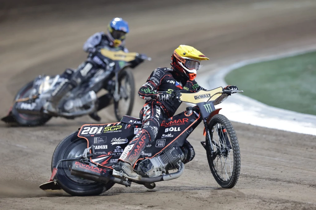 Jan Kvech, a za nim Maciej Janowski Dwóch żużlowców w kolorowych kombinezonach ściga się na torze żużlowym, motocykle pochylone w zakręcie, widoczny kurz unoszący się nad powierzchnią toru.