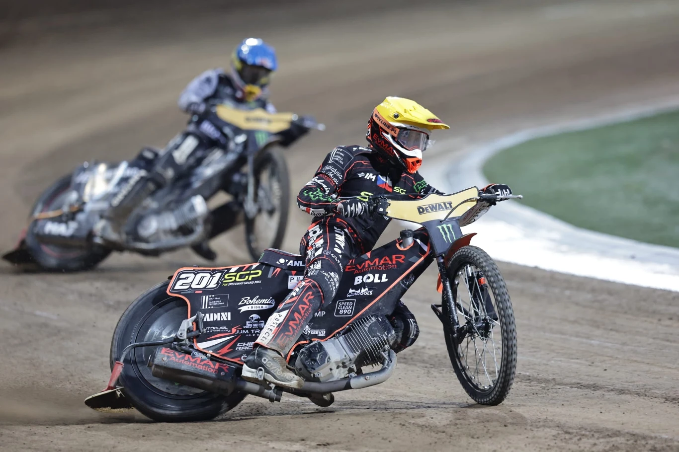 Jan Kvech, a za nim Maciej Janowski Dwóch żużlowców w kolorowych kombinezonach ściga się na torze żużlowym, motocykle pochylone w zakręcie, widoczny kurz unoszący się nad powierzchnią toru.