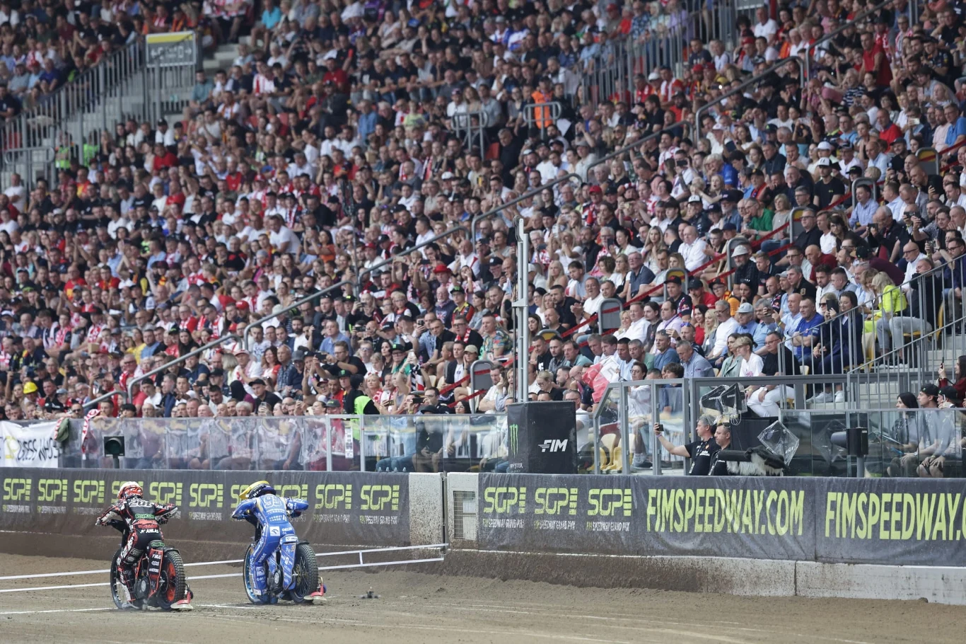 Jan Kvech oraz Bartosz Zmarzlik, a w tle trybuny Stadionu Olimpijskiego we Wrocławiu Dwóch zawodników na motocyklach żużlowych przygotowuje się do startu na torze, a trybuny wypełnione są liczną publicznością kibicującą wydarzeniu sportowemu.