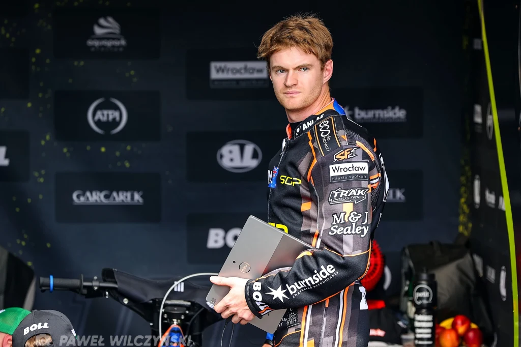 Brady Kurtz Młody mężczyzna w kombinezonie wyścigowym trzymający laptop na tle stołu z owocami oraz elementami technicznymi, za nim widoczne logotypy sponsorów i czarny motocykl sportowy.