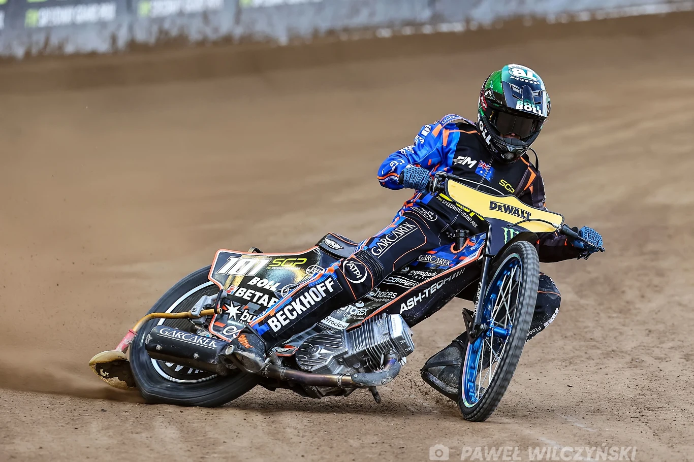 Zawodnik żużlowy w sportowym kombinezonie i kasku pędzi motocyklem po torze żużlowym, wykonując dynamiczny zakręt na sypkiej nawierzchni. Motocykl pokryty jest licznymi logotypami sponsorów.