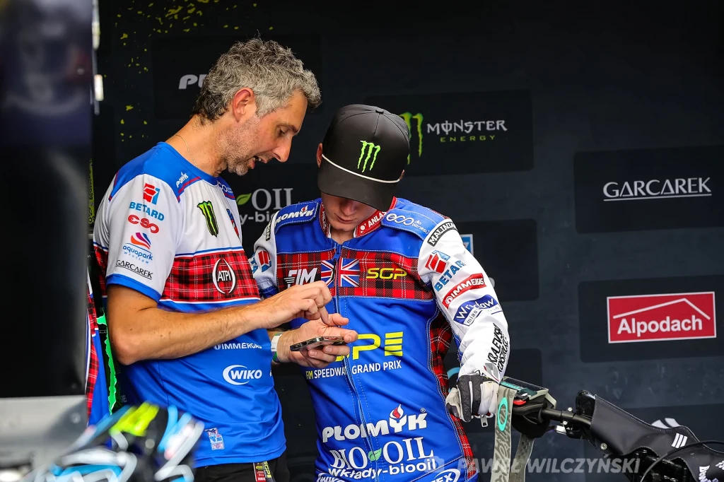 Dwóch mężczyzn w ubraniach z logotypami sponsorów omawia coś na ekranie telefonu komórkowego, jeden z nich nosi strój sportowy i czapkę z daszkiem, obok widoczny motocykl żużlowy.