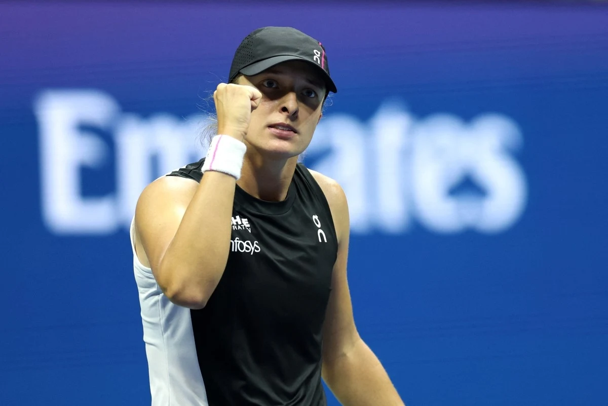 Tenisistka w czarnej czapce i stroju sportowym zaciśnięta pięść w geście triumfu na korcie tenisowym, niebieskie tło z rozmazanym napisem.