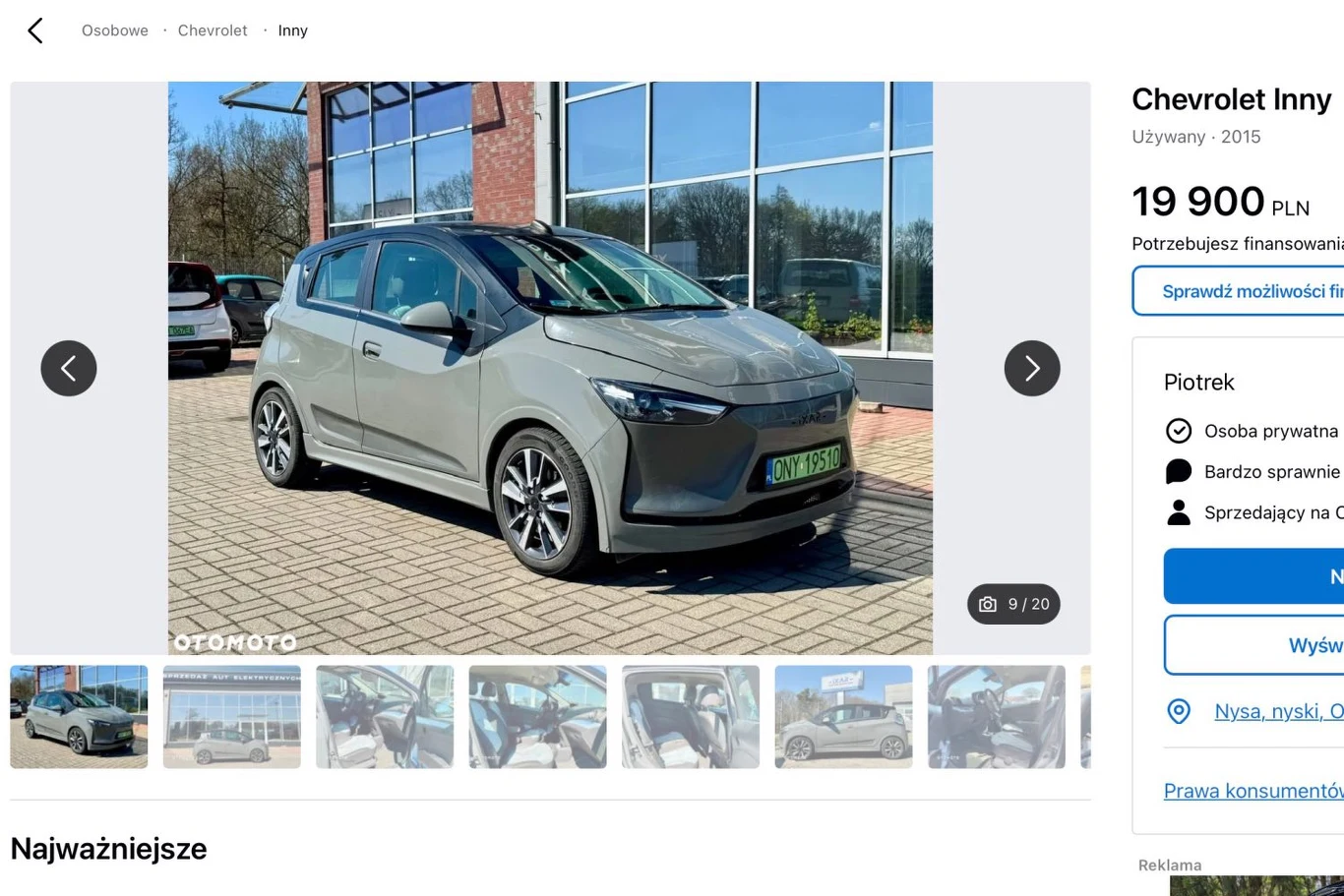 Srebrny samochód osobowy Chevrolet zaparkowany na chodniku przed nowoczesnym budynkiem z dużymi oknami, w tle widoczne drzewa i niebo.