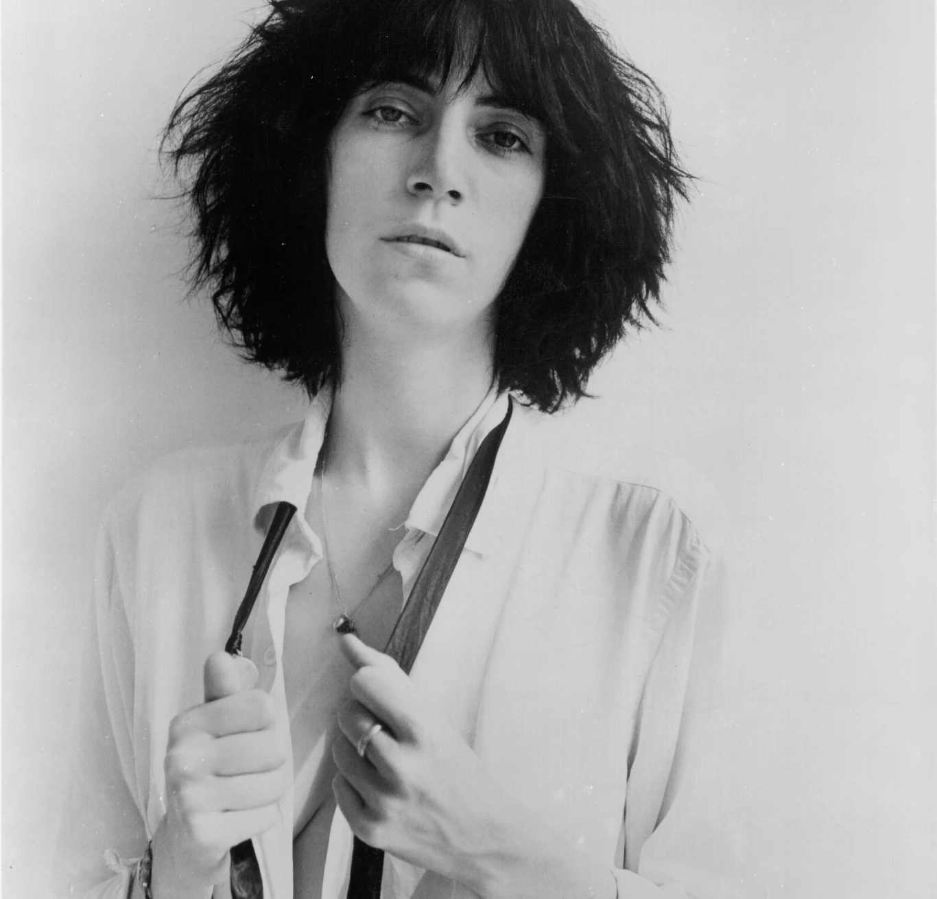 Patti Smith w połowie lat 70. Kobieta o ciemnych, potarganych włosach patrzy prosto w obiektyw, trzymając rozpiętą koszulę i szelki na tle jasnej, jednolitej ściany.
