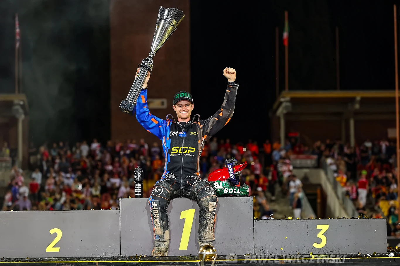 Zwycięzca turnieju żużlowego trzymający puchar nad głową, stojący na najwyższym stopniu podium z podniesioną ręką w geście triumfu, wokół widoczni kibice na trybunach.