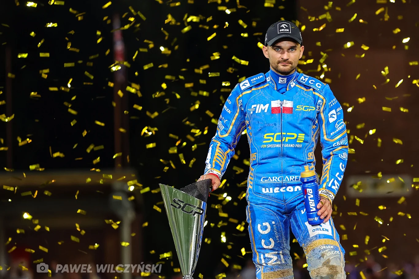 Niepocieszony Bartosz Zmarzlik Żużlowiec w niebieskim kombinezonie sportowym trzymający puchar, wokół niego unoszą się złote konfetti. Mężczyzna ma poważny wyraz twarzy i stoi na tle rozświetlonej areny.