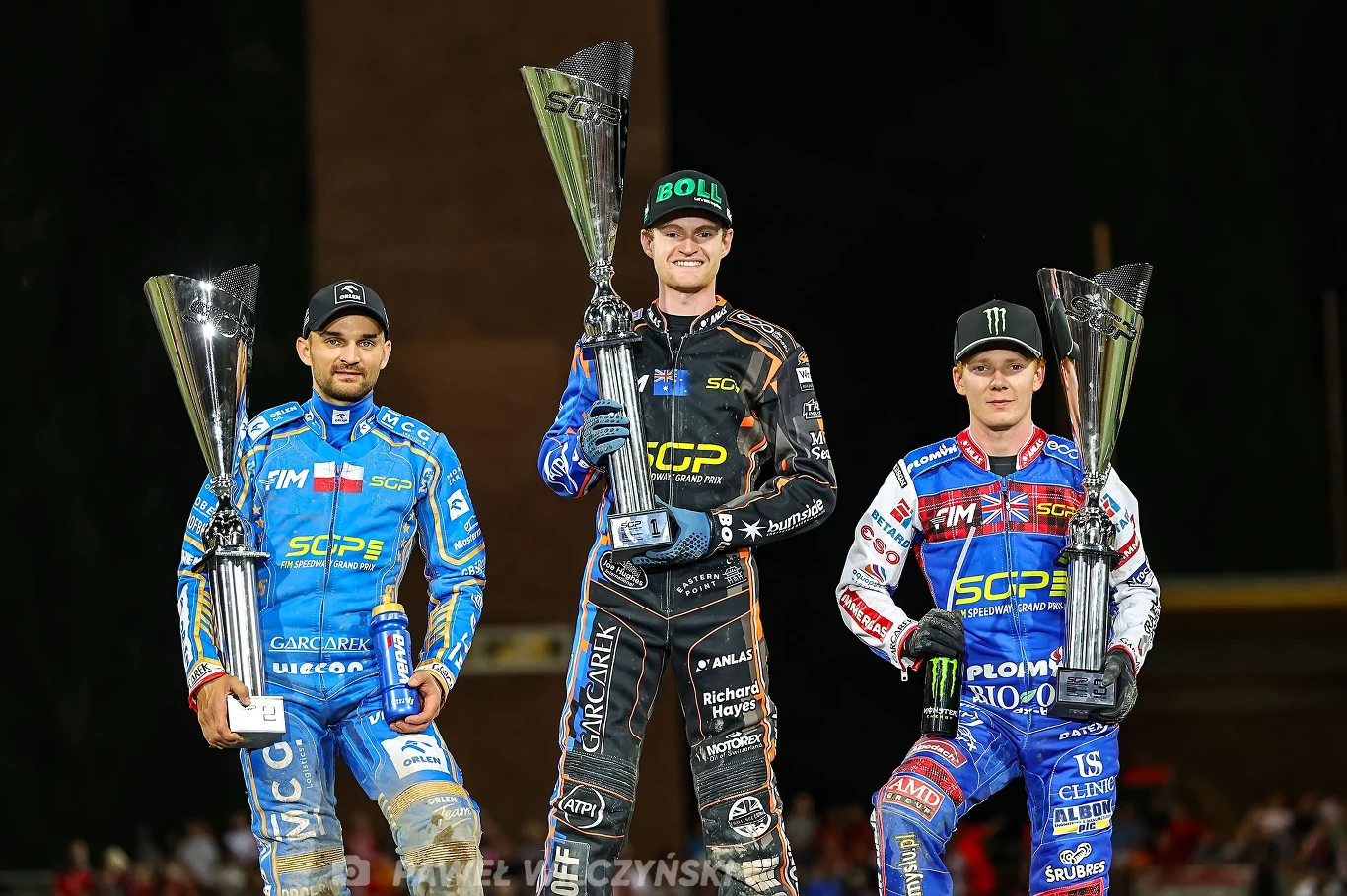 Podium Grand Prix Polski we Wrocławiu. Zmarzlik, Kurtz i Bewley Trzech żużlowców w kolorowych kombinezonach stoi na podium z dużymi pucharami, wszyscy uśmiechnięci, centralny zawodnik wyższy od pozostałych, tło zamazane, skupienie na triumfatorach zawodów.