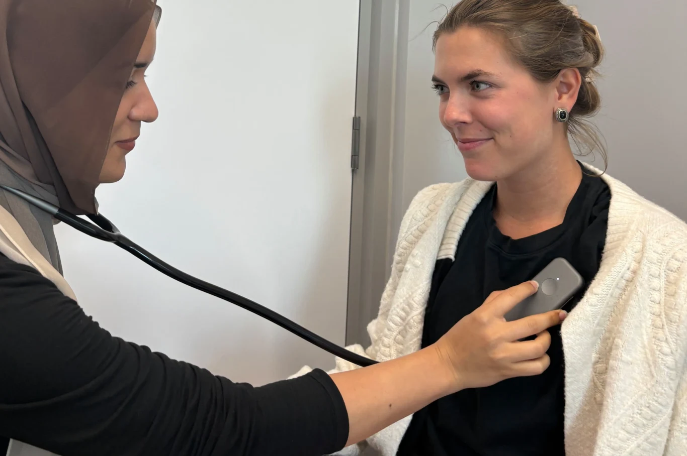 Stetoskop doczekał się cyfrowej rewolucji. Diagnoza serca w 15 sekund Lekarka w chuście bada stetoskopem kobietę o blond włosach, która siedzi spokojnie i lekko się uśmiecha, obie znajdują się w jasnym pomieszczeniu o neutralnym tle.