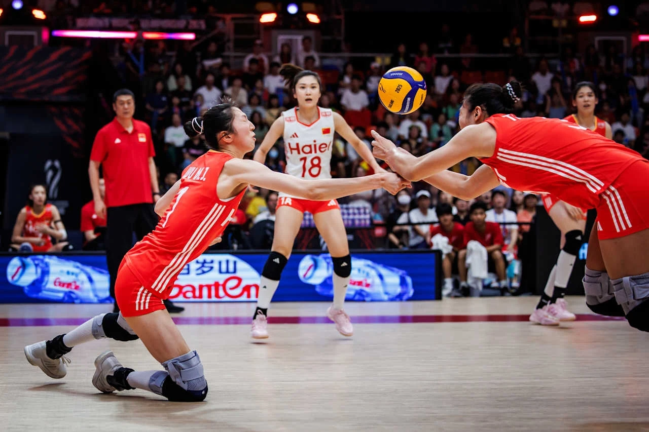 Siatkarki reprezentacji Chin Zawodniczki drużyny siatkarskiej w czerwonych strojach podczas dynamicznej akcji odbicia piłki, na tle publiczności i sędziów w hali sportowej.