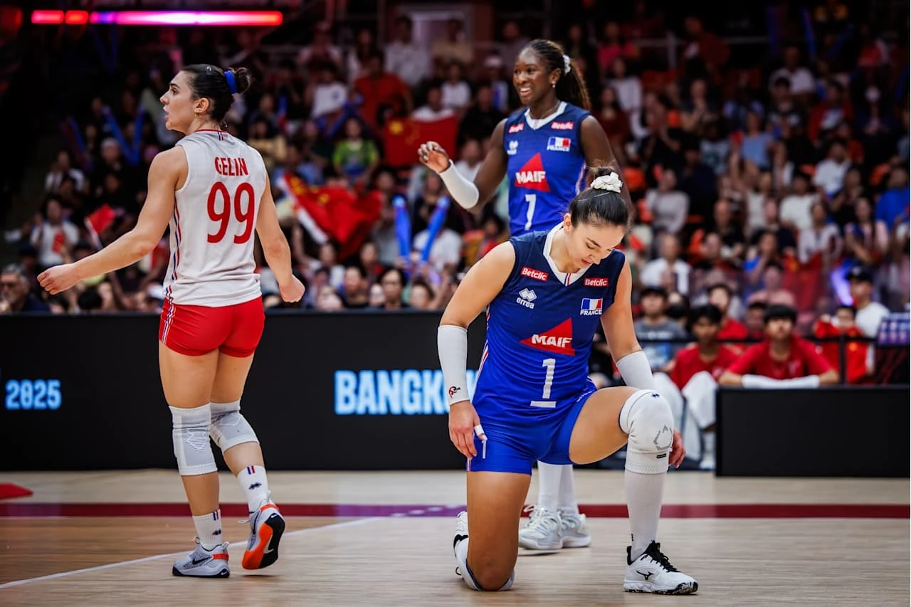 Siatkarki reprezentacji Francji Zawodniczka w niebieskim stroju klęczy na kolanie na środku hali sportowej, wokół niej widoczne są inne siatkarki oraz tłum kibiców na trybunach.
