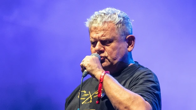 Mężczyzna o siwych włosach śpiewa do mikrofonu na scenie, nosi czarną koszulkę z żółtym napisem i czerwoną opaskę na nadgarstku, w tle widać fioletowe światła sceniczne.