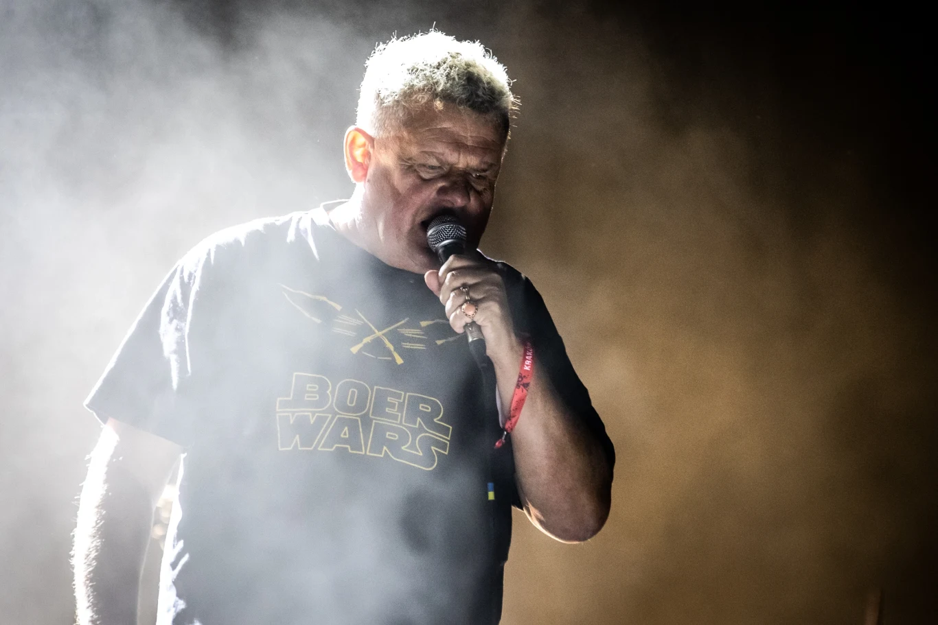 Kazik Staszewski (Kult) Mężczyzna z siwymi włosami śpiewający do mikrofonu, ubrany w czarną koszulkę z napisem 'Boer Wars', stojący wśród intensywnego oświetlenia i dymu scenicznego.