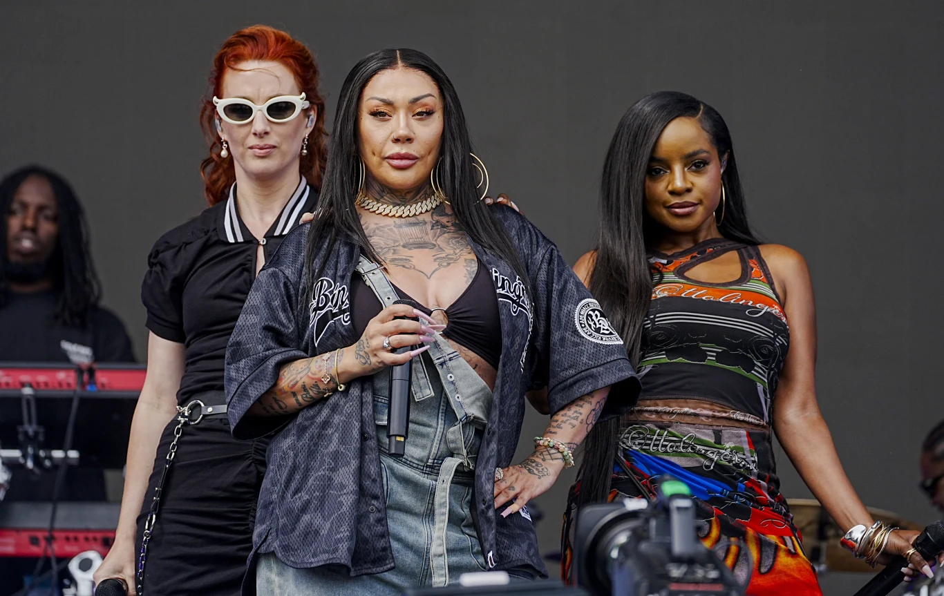 Zespół Sugababes podczas BBC Radio 1's Big Weekend w 2025 r. Trzy kobiety pozujące na scenie, centralna z mikrofonem w ręku, ubrane stylowo i wyraziście, w tle członkowie zespołu oraz sprzęt muzyczny.