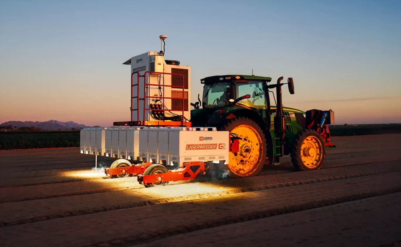 Chwasty nie mają szans z laserami i sztuczną inteligencją. Zdrowszy sposób na rolnictwo Nowoczesny traktor ciągnący zautomatyzowaną maszynę rolniczą oświetloną reflektorami, pracującą na polu o zmierzchu.