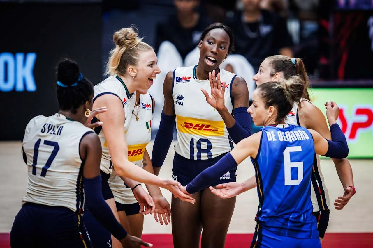Grupa zawodniczek drużyny siatkarskiej cieszy się po zdobytym punkcie, ubrane w sportowe stroje, numerowane koszulki z nazwiskami, emocje i radość na twarzach, w tle rozmyte sylwetki kibiców.