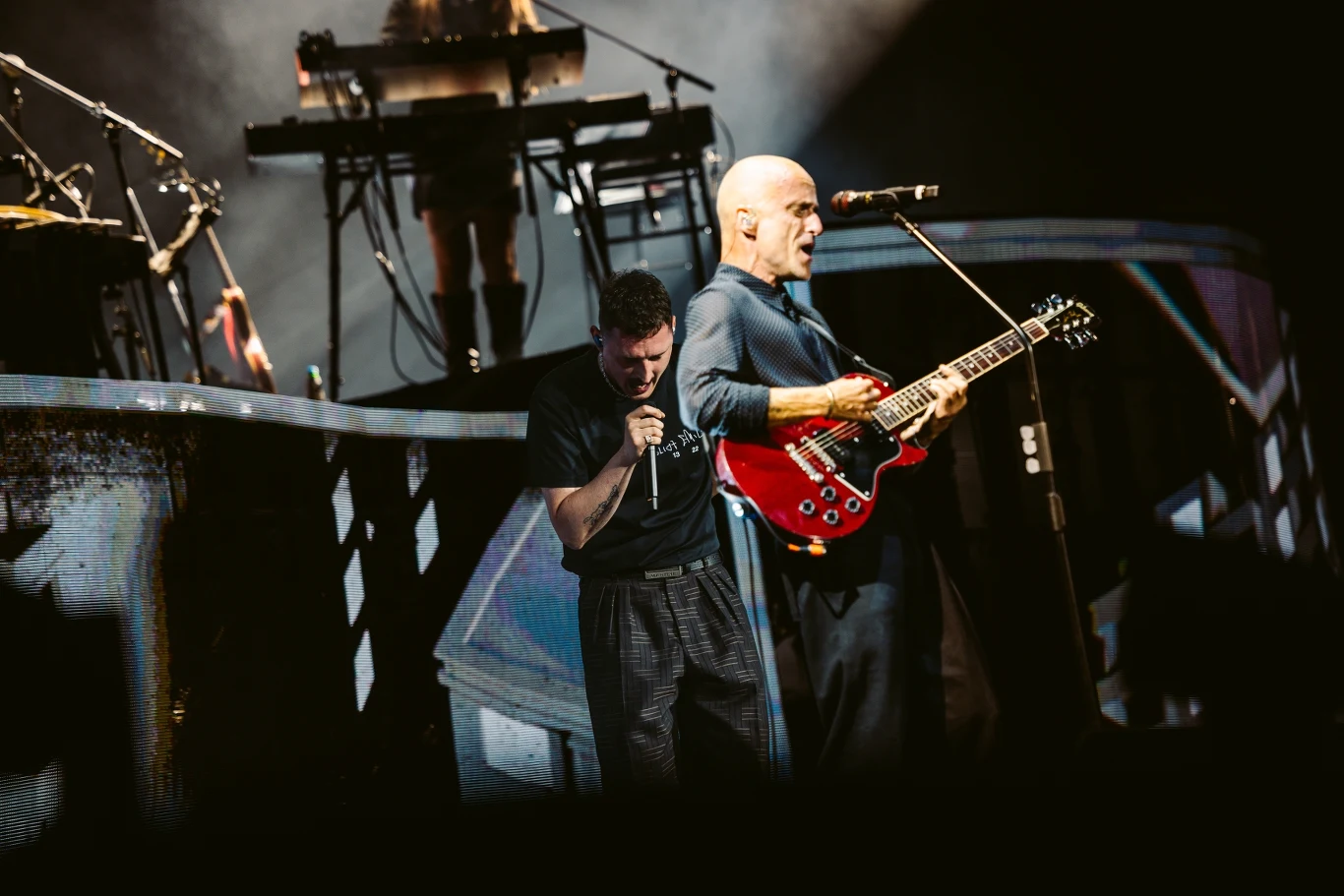 Dwóch muzyków występujących na scenie, jeden z gitarą elektryczną i mikrofonem śpiewa, drugi trzyma mikrofon i śpiewa, w tle widoczny jest klawiszowiec z instrumentami na podwyższeniu, wokół efektowne oświetlenie sceny.