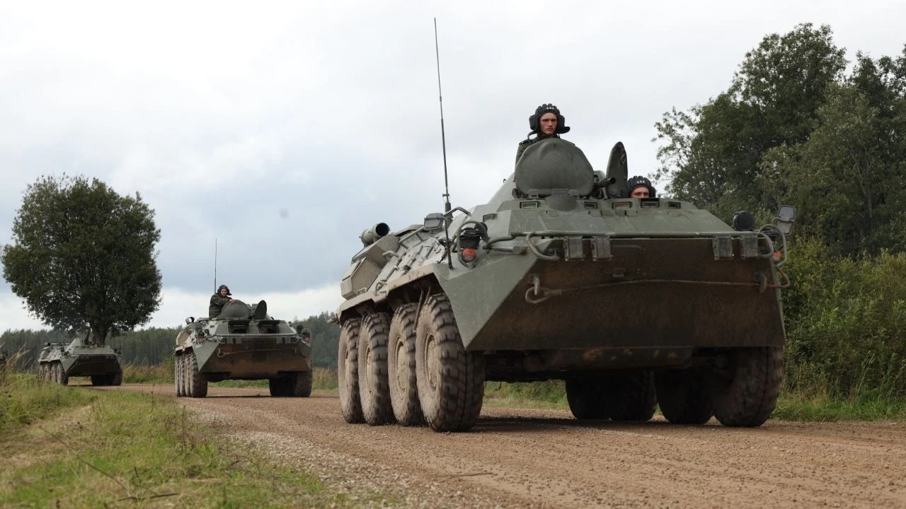 Manewry wojskowe krajów OUBZ na Białorusi Konwój wojskowych transporterów opancerzonych poruszający się po nieutwardzonej drodze w otoczeniu zieleni, żołnierze widoczni w wieżyczkach pojazdów, niebo zachmurzone, atmosfera spokojna.