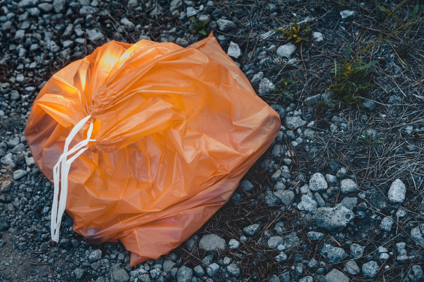Pomarańczowy worek wśród śmieci. Coś się właśnie zmieniło (zdj. ilustracyjne) Pomarańczowy worek na śmieci leżący na nierównej, kamienistej ziemi, otoczony suchą trawą i drobnymi kamieniami.