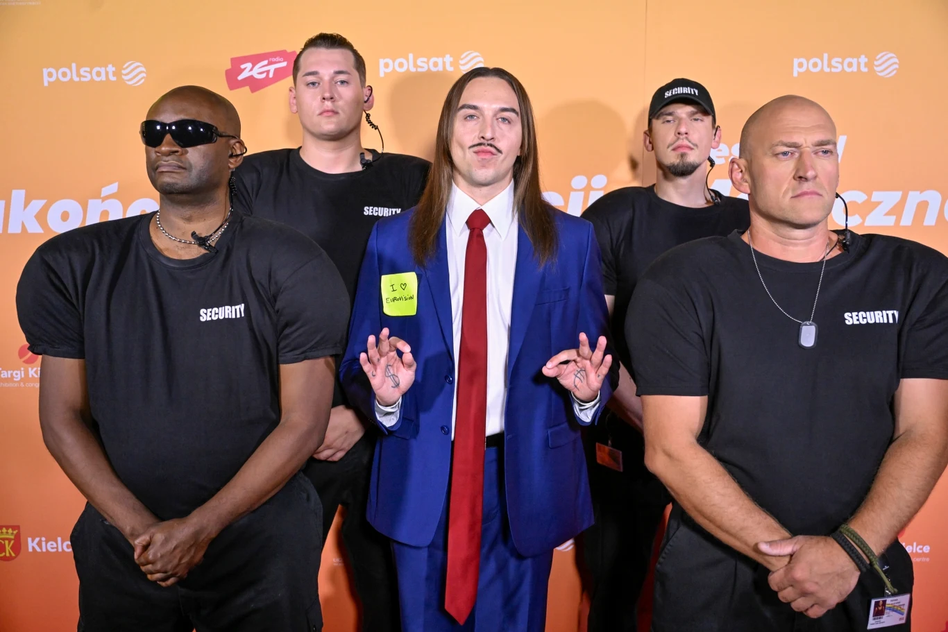 Tommy Cash i jego ekipa w Kielcach Pięciu mężczyzn pozujących na tle pomarańczowej ścianki z logo stacji telewizyjnej, czterech z nich ubranych w czarne koszulki z napisem 'Security', a jeden w niebieskim garniturze z czerwoną krawatą i identyfikatorem.