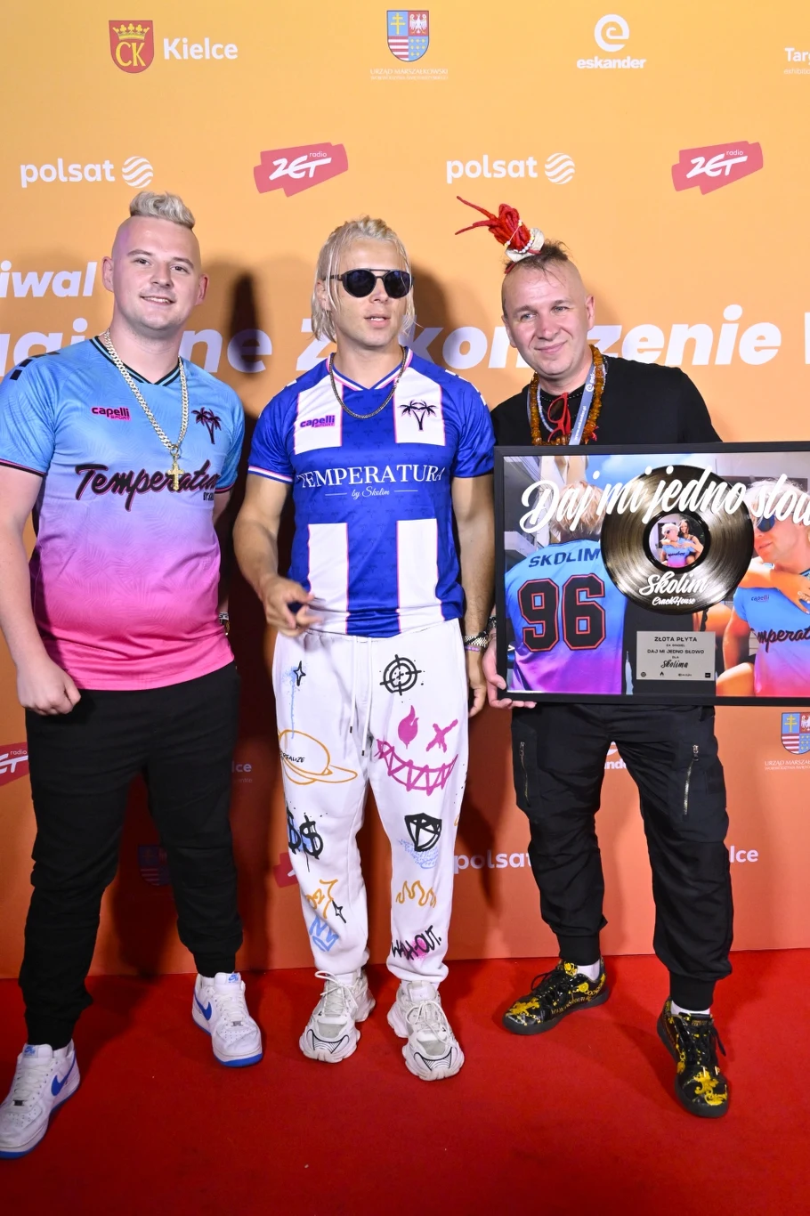 Skolim i jego zespół Trzech mężczyzn stojących na czerwonym dywanie na tle ścianki z logotypami sponsora, jeden z nich trzyma tablicę pamiątkową z napisem, a pozostali pozują do zdjęcia w kolorowych ubraniach.