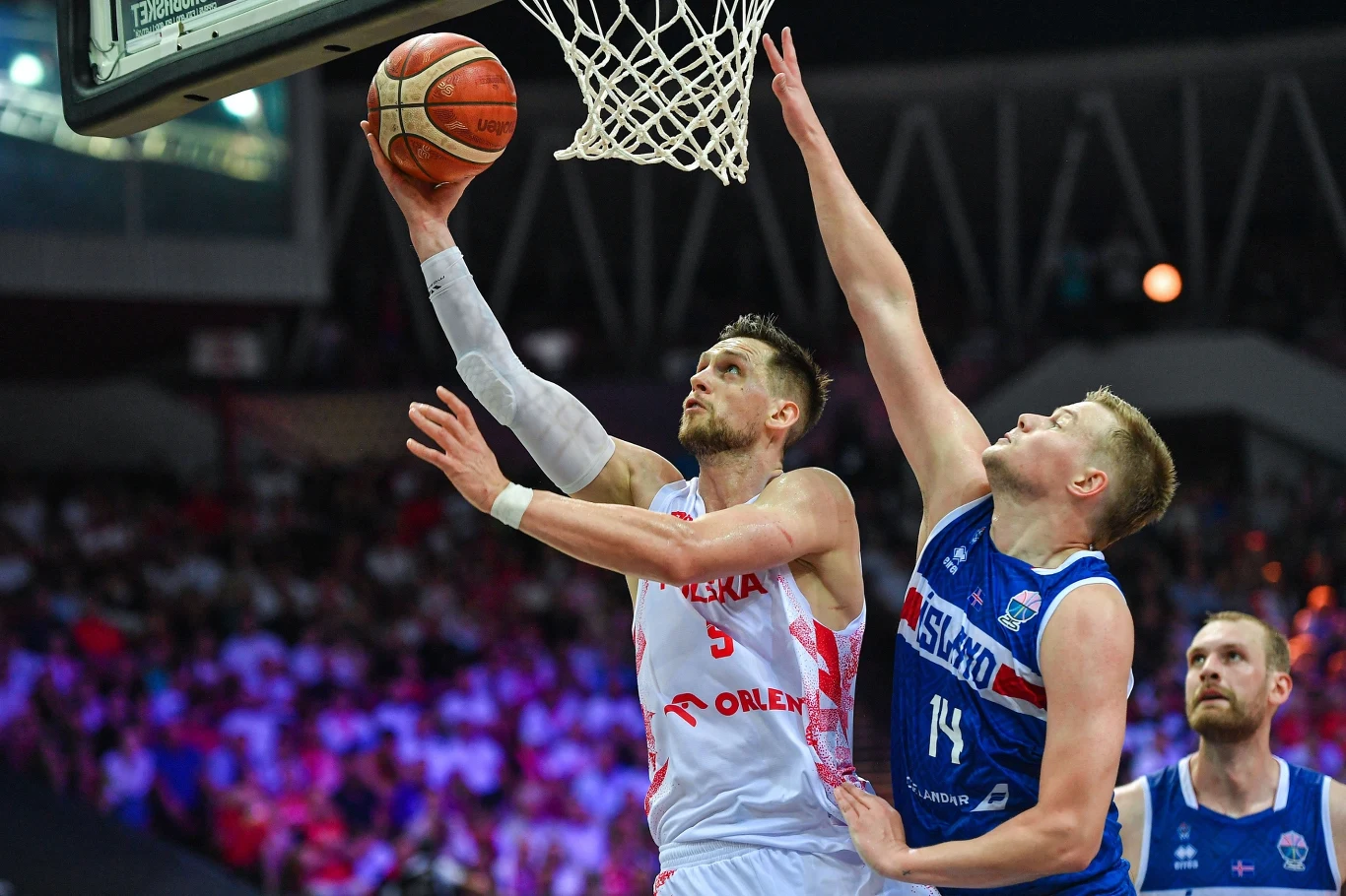 Mateusz Ponitka łatwo uciekał rywalom Zawodnik w białym stroju drużyny Polski wykonuje dynamiczny rzut do kosza, broniony przez zawodnika w niebieskim stroju, w tle widoczna publiczność oraz elementy hali sportowej.