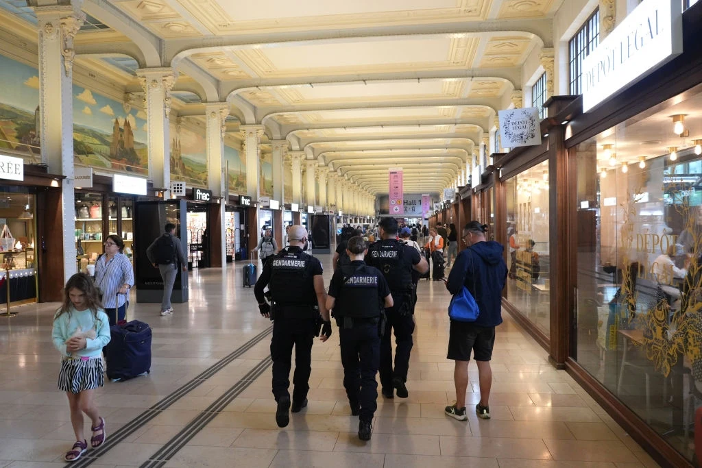 Policja patrolująca paryski dworzec Gare de Lyon (zdjęcie ilustracyjne) Korytarz dworca lub galerii handlowej z wysokim ozdobnym sufitem, patrol służb mundurowych idący przez środek, po bokach liczne sklepy, kilka osób przechadzających się i dziecko z walizką.