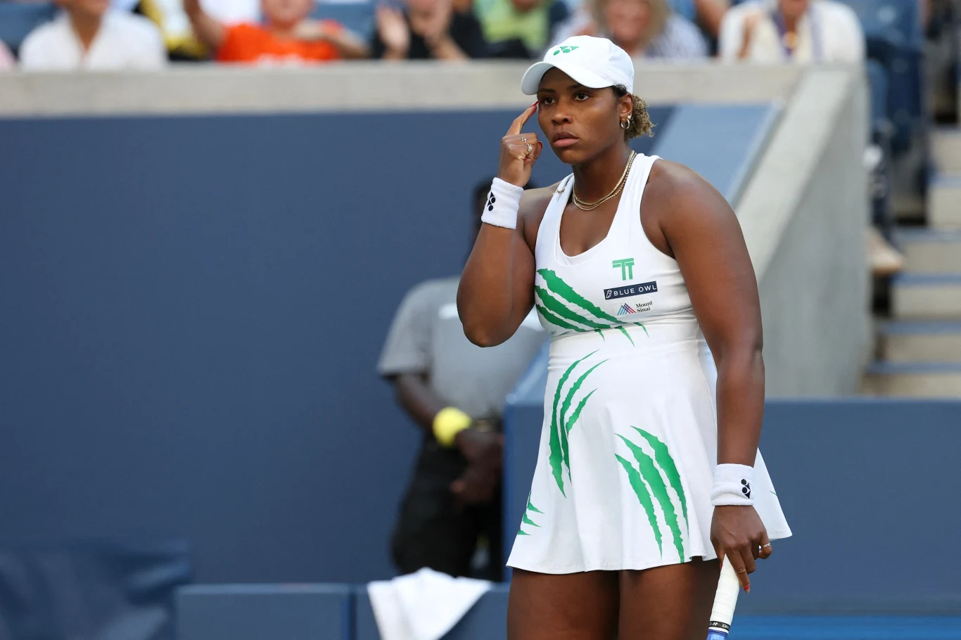 Taylor Townsend Tenisistka w białym stroju sportowym z zielonymi akcentami na korcie tenisowym, skupiona przed wykonaniem serwisu, trzyma rakietę w lewej ręce, za nią rozmyte sylwetki widzów na trybunach.