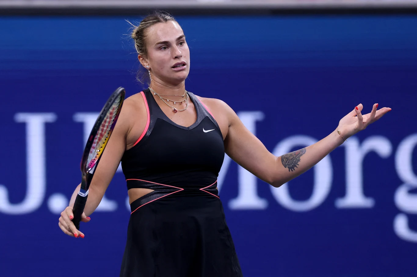 Aryna Sabalenka, US Open 2025 Tenisistka w czarnej sportowej sukience z rakietą w ręce, wyrażająca frustrację lub zaskoczenie na tle niebieskiego kortu z widocznym fragmentem reklamy turnieju.